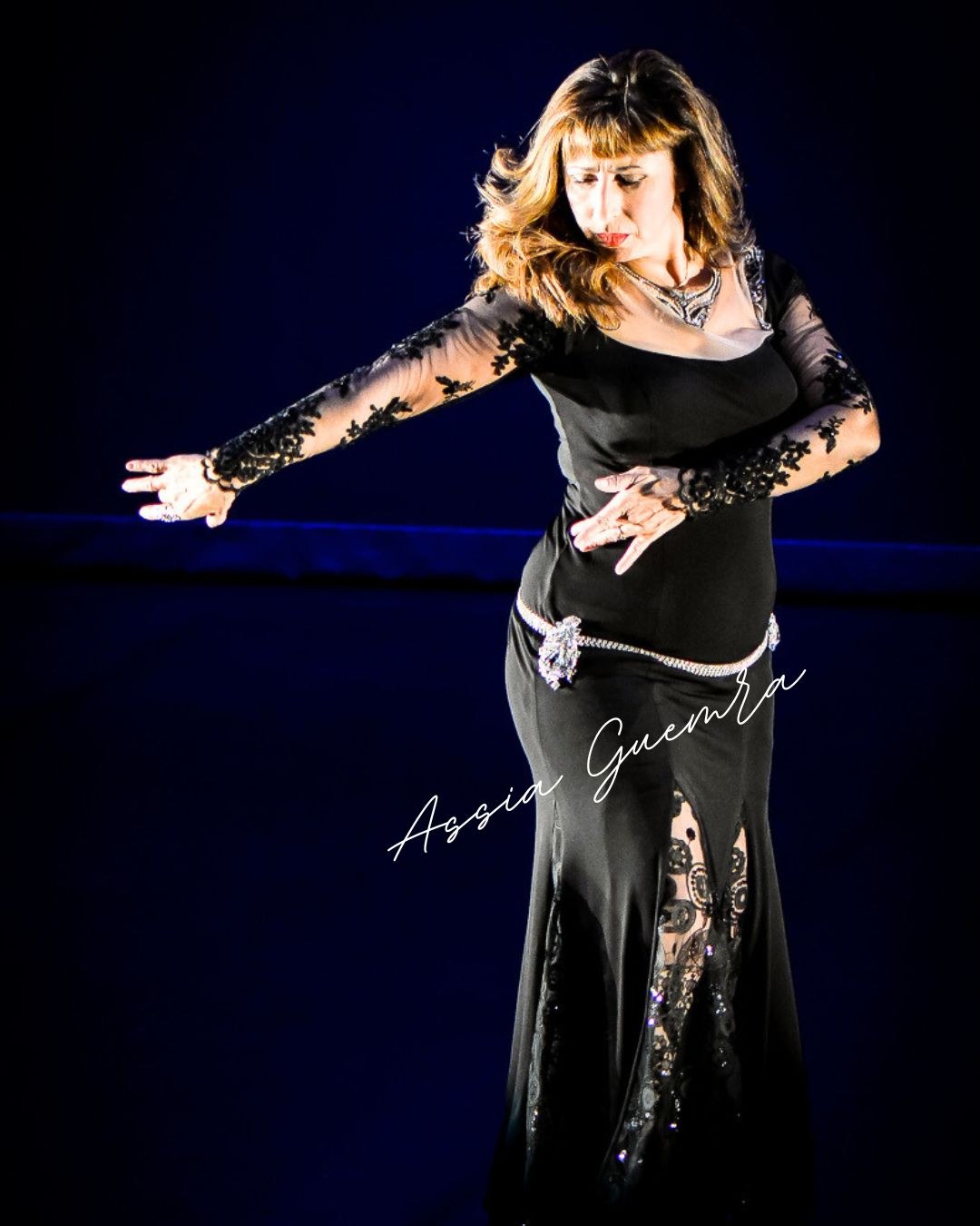 Assia en robe de scène noire brodée, un bras tendu sur le côté, sur une scène au fond bleu nuit