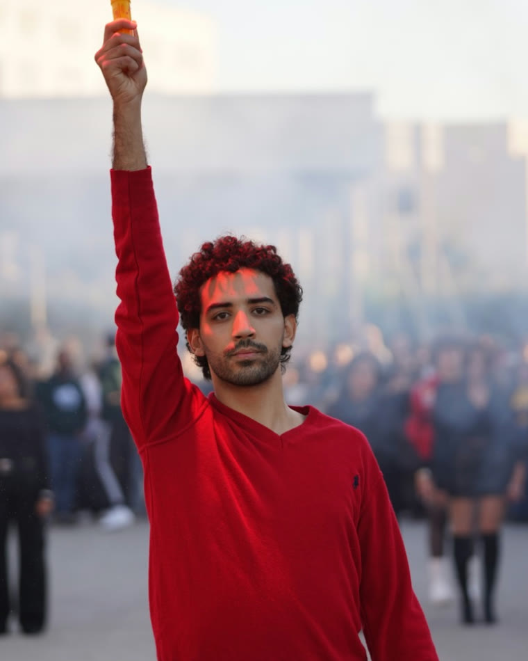 Elyes en pull rouge, bras levé, devant un groupe de danseurs en extérieur