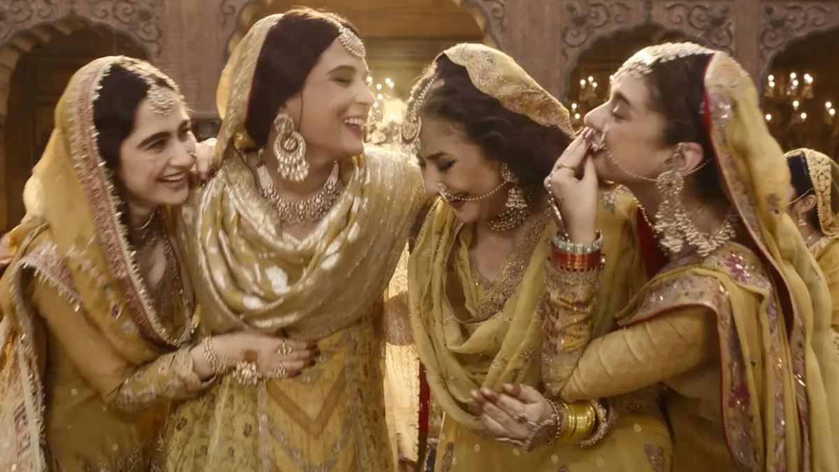 Quatre danseuses en tenues jaune safran, riant ensemble, dans un palais éclairé de lustres